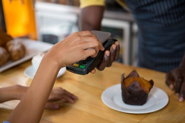 A person holding a smartphone displaying the Apple Pay interface, with multiple bank cards added to the wallet. The background shows a coffee shop counter with a visible contactless payment terminal.