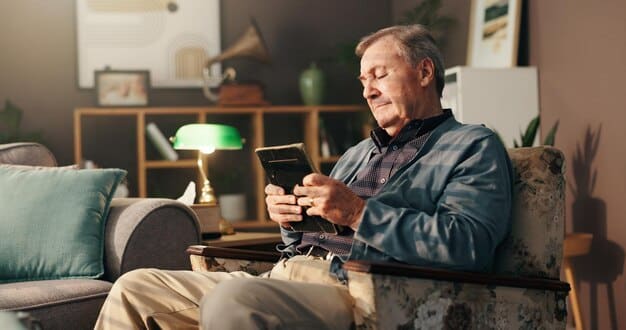 A senior man sitting comfortably in his living room, using a tablet to pay his bills through a mobile payment app. The room is cozy, with soft lighting and a cup of tea on the table next to him.